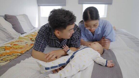 年轻的爸爸和妈妈在睡床上玩着一个5个月大的可爱的女婴躺在卧室的床上亚洲家庭在早上一起度过周末时光感受幸福的时刻积极的情绪