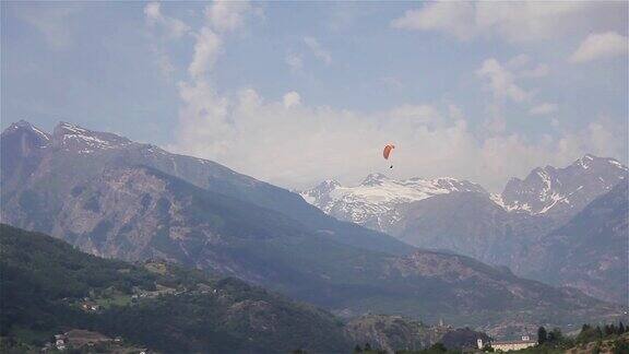 滑翔伞在遥远的山地景观一个滑翔伞飞行使用橙色伞漂浮下降缓慢盘旋在树太阳雪峰夏天视频观看山谷空中体育旅游