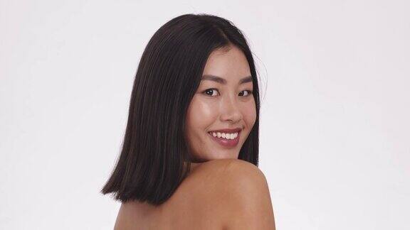 顽皮的亚洲女人转向镜头和微笑抚摸她完美的肩膀皮肤慢镜头