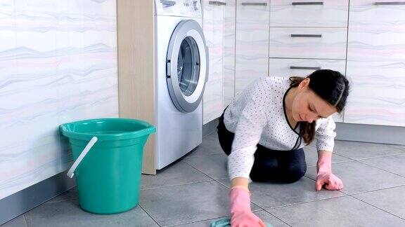 戴着粉色橡胶手套的疲惫的女人用一块布洗厨房的地板看着镜头地板上铺着灰色瓷砖