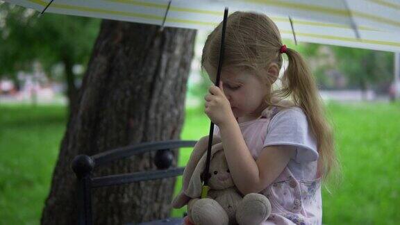 一个小女孩带着一只玩具兔子坐在一把伞下的长椅上
