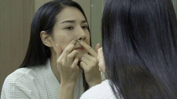 年轻美丽的亚洲女人看着她的脸皮肤挤在浴室镜子上的青春痘