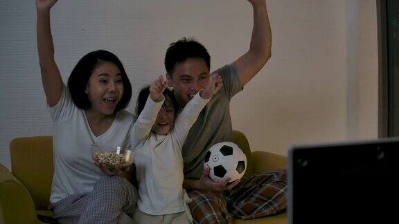 亚洲家庭坐在沙发上一起看电视足球赛为胜利和胜利而欢欣鼓舞