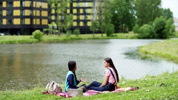 两个大学女生坐在公园池塘边的毯子上聊天放松的学生亚洲和高加索人课后在自然中聊天周围的书籍笔记本电脑和背包