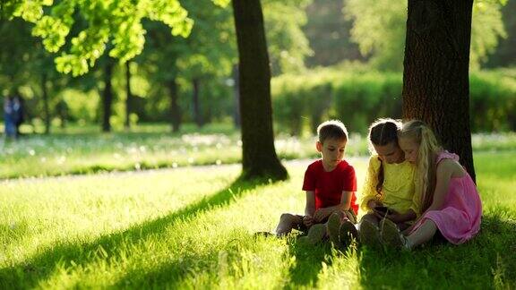 三个孩子两个女孩和一个男孩坐在公园树下的草地上在阳光明媚的夏日用智能手机追踪拍摄