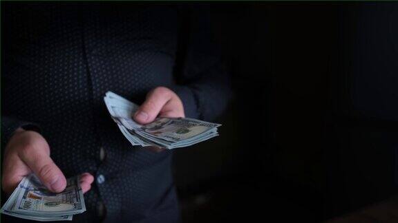 一名身穿黑衬衫、手里拿着钱的男子站在黑暗中从另一个人手里拿了一包100美元的钞票