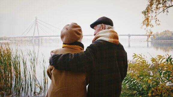 秋日老丈夫拥抱着老妻子微笑着和她说话站在河岸上看着桥