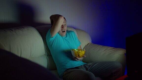 一个男人看起来很震惊深夜坐在沙发上吃着一碗薯条看电视