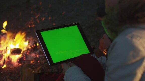 晚上女孩在篝火旁拿着一台绿色屏幕的平板电脑