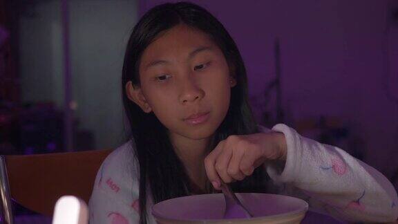 可爱的亚洲女孩晚餐吃米饭汤在家用LED灯通过智能手机看VDO剪辑