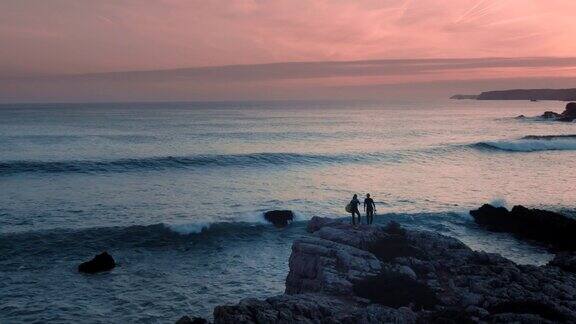 美丽的航拍两名女性冲浪者站在悬崖岩石的边缘观看日落时的海浪