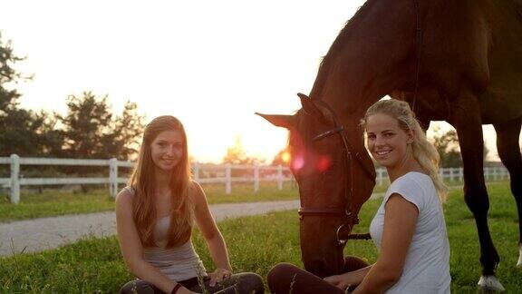 慢镜头:两个女孩坐在草地上在日落时分和马一起闲逛