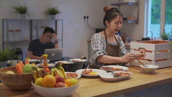 亚洲华人男子在家里用笔记本电脑工作而家庭主妇在厨房准备早餐