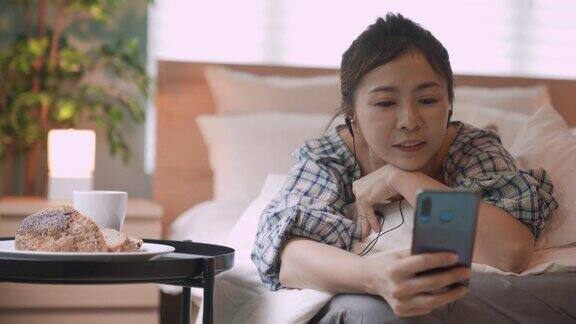 亚洲中国女人躺在床上用手机在卧室吃早餐