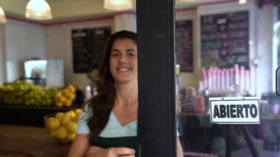 一个漂亮的果汁酒吧的老板走到门口在窗户上放了一个打开的标志看着镜头微笑着