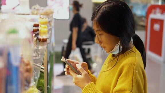 亚洲女孩戴着口罩在美食广场购买零食和饮料然后通过手机二维码支付新常态的生活理念