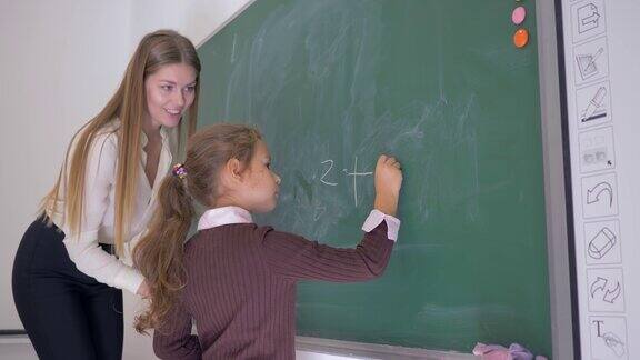 数学课上一个女学生在女老师的帮助下用一支粉笔在黑板上写下一个例子