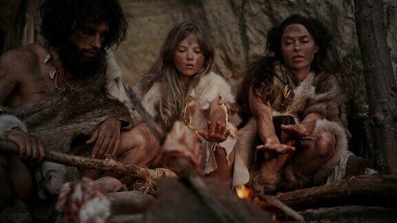 穿着兽皮的史前狩猎采集者部落晚上住在洞穴里尼安德特人或智人家庭试图在篝火取暖手在火上烹饪食物