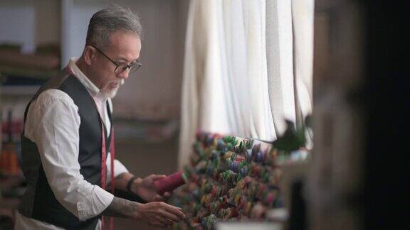 面部留胡茬的亚洲华人成熟男子裁缝在工作室检查他的织物服装
