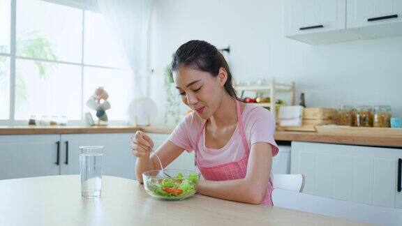 穿着围裙的亚洲美女在家里的厨房里吃着蔬菜沙拉年轻美丽的女孩感到快乐享受早餐与蔬菜健康的食物和清洁的水以饮食的健康在家里
