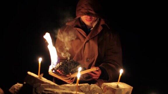 一个戴着兜帽披着斗篷的男人正在读一本热乎乎的书地上还放着蜡烛