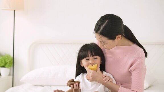 幸福的家庭年轻的妈妈保姆和可爱的小女儿一起坐在床上吃甜甜圈