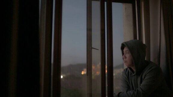 亚洲华人中年男子看窗外从他的家在日落与严肃的面部表情与帽衫
