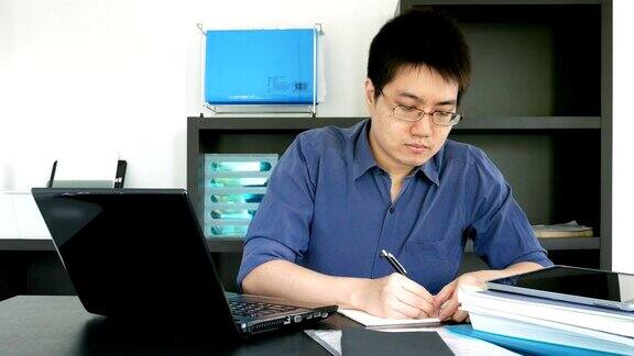 戴眼镜的亚洲商人用笔记本电脑工作并在家庭办公室的笔记本上写作