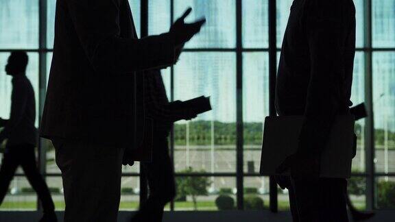 两个商人的剪影拿着笔记本和记事本在办公室大厅互相握手和交谈人们在后面走过