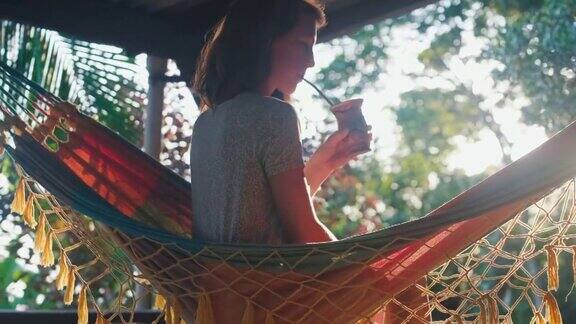 女人喝的伴侣年轻的白人妇女坐在吊床上在日出时喝着葫芦里的酒在花园里喝花草茶的女人的肖像
