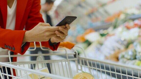 在超市:女人用智能手机靠在购物车上在大商场里用手机上网的女人专注于手持手机