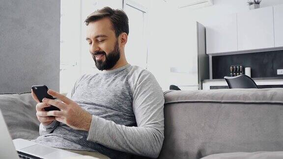 快乐的大胡子男人一边坐在沙发上用笔记本电脑一边玩智能手机