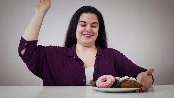 微笑的白人肥胖女孩坐在糖果和卷尺前的肖像胖女人把卷尺移到一边拿起装有甜点的盘子肥胖、饮食、自控力