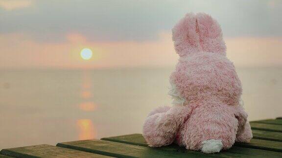 粉红色的玩具兔子坐在码头上看着冉冉升起的太阳