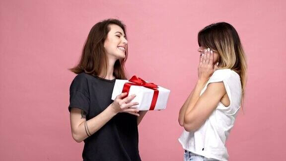 快乐的年轻女孩给她的女性朋友礼物盒和拥抱她孤立的粉红色背景