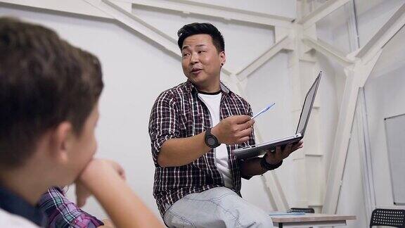 韩国老师坐在桌子上手里拿着笔记本电脑给学生上课小学生认真听讲向老师提出重要问题