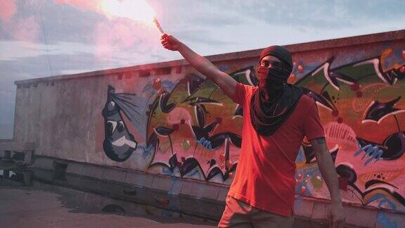 一个穿着巴拉克拉瓦帽的年轻人在屋顶上挥舞着红色的信号弹背景是涂鸦慢动作
