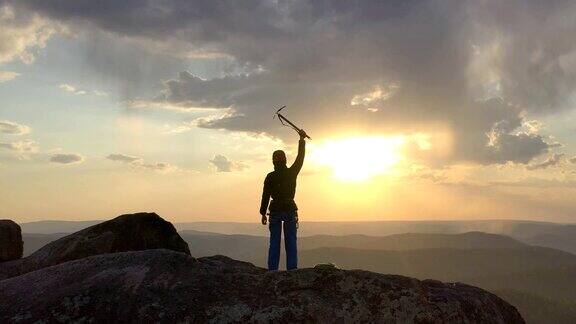 日落时分一名男子站在一座石山的山顶上手拿冰镐开心地挥舞着双手