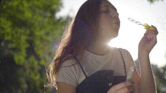 一个十几岁的女孩在公园的阳光下对着摄像机吹肥皂泡