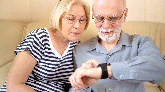 一对愉快、好看的老年夫妇坐在家里的沙发上使用智能手表浏览阅读
