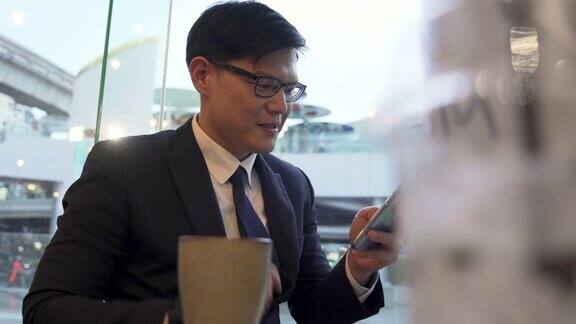 亚洲商人办公室职员坐在咖啡店的窗口边喝咖啡使用智能手机和互联网在线工作公司业务或短信商业和技术概念