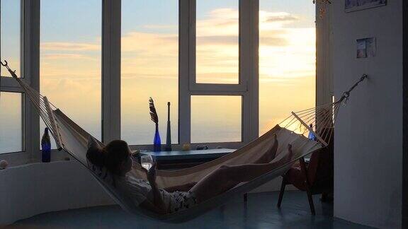 年轻女性喝葡萄酒在家中对着全景窗户在吊床日落