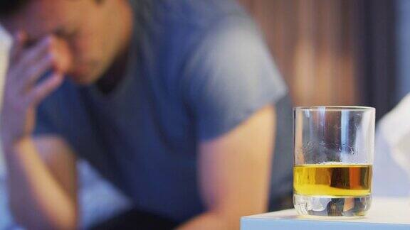 一个沮丧的男人穿着睡衣坐在床边床头柜上放着一杯威士忌
