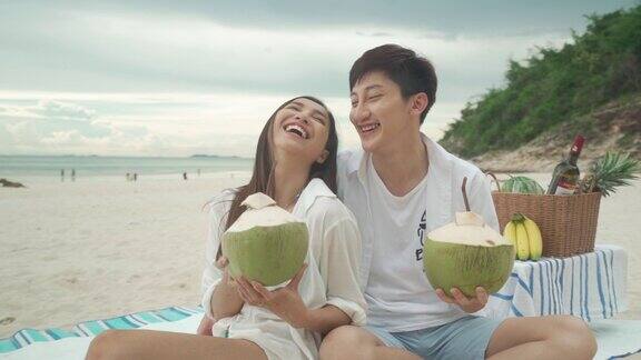 亚洲年轻夫妇坐在野餐毯上在海滩和海边喝椰子汁背景是热带水果夏季、假期、假期和快乐的泰国人概念