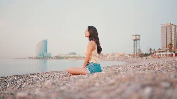 美丽的棕色头发的女人长头发坐在海滩上的海景背景华丽的女孩穿着上衣和牛仔短裤慢动作