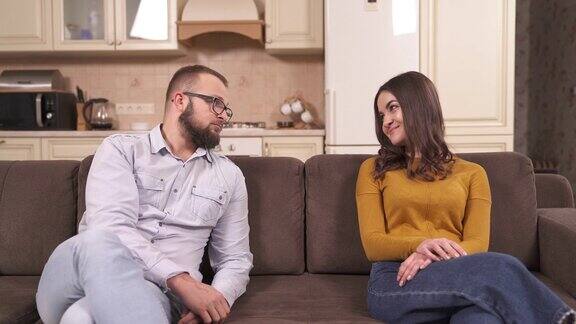 一个戴眼镜的大胡子男人和一个漂亮的黑发女人坐在一间单间公寓角落的沙发上;他们拍着手看着对方笑