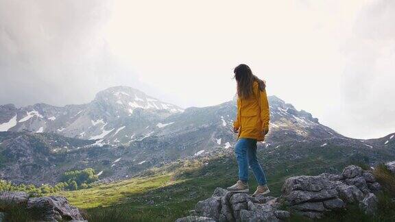 年轻快乐的女人穿着黄色雨衣在岩石上行走背景是美丽的山脉慢镜头