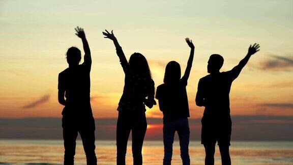 背影剪影四个不同的年轻朋友站在海边的海滩上看着美丽的橙色夕阳在水面上挥手感觉快乐和自由