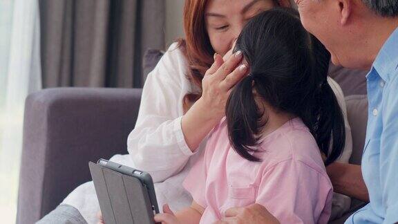 亚洲的祖父母和孙女在家里使用平板电脑大四的中国爷爷奶奶开心的花家人放松与年轻的女孩查看社交媒体躺在客厅的沙发上缓慢的运动