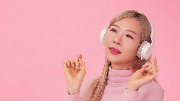 韩国女孩的肖像听音乐通过耳机和舞蹈孤立在工作室的粉红色背景